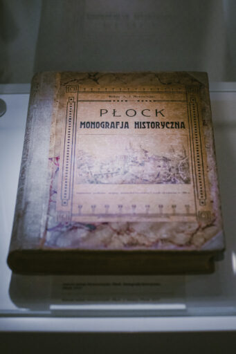 Galeria płocczan XX wieku. W gablocie leży stara książka. Ma zdobioną okładkę z napisem "Płock. Monografia historyczna".