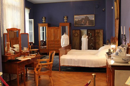 Secesyjna sypialnia z Muzeum Mazowieckim w Płocku, umeblowana łóżkiem zasłanym białą pościelą, dużą drewnianą szafą, po lewej stronie stoli toaletka z lustrem, przed nią krzesło. Przy łóżku stoją manekiny ubrane w stroje nocne z epoki secesyjnej.