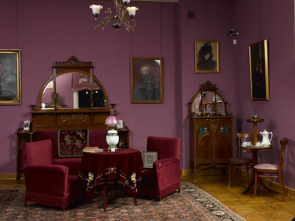 Secesyjny buduar - pomieszczenie o fioletowych ścianach, na podłodze parkiet. Wypełniają je staromodne meble z ciemnego drewna, obite czerwonym pluszem fotele, stolik, na stoliku wazon.