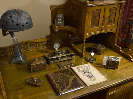 Biurko w secesyjnym gabinecie pana domu w muzeum, na nim staromodny komplet przyborów do pisania, kałamarz, osuszacz do papieru, wszystko ozdobione kwiatowymi motwami. Po lewej stronie na biurku metalowa, ozdobna lampa.