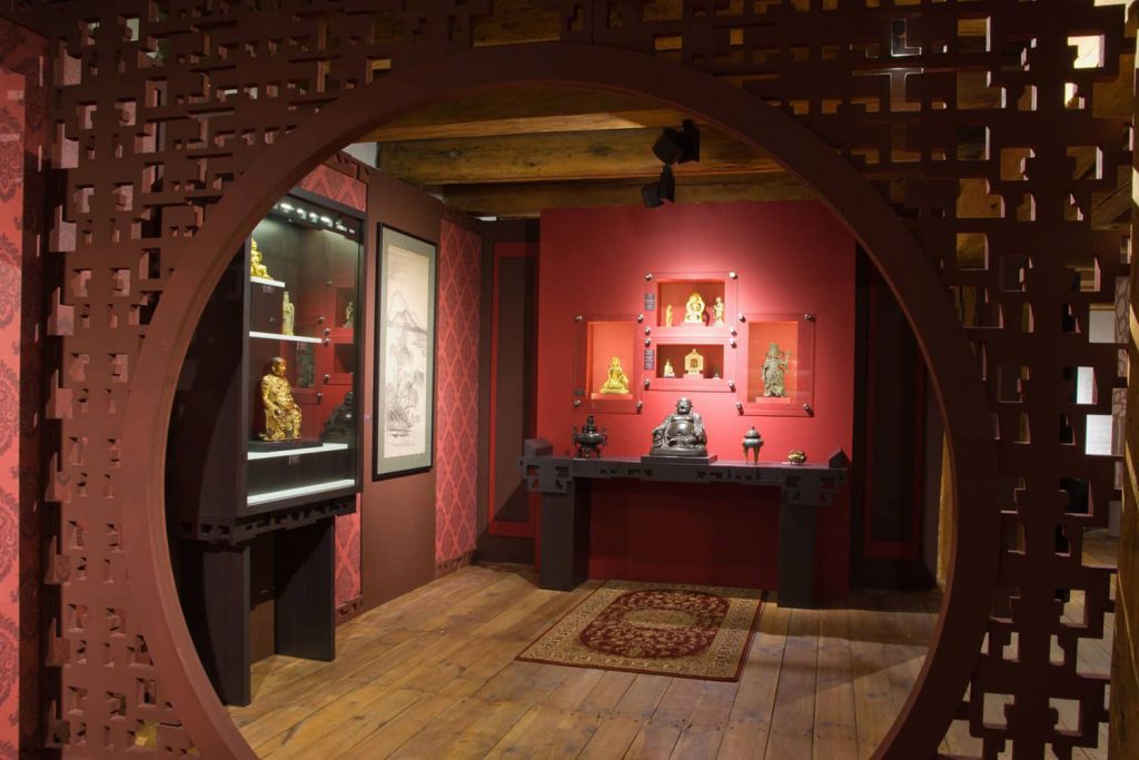 Wystawa "Sztuka Dalekiego Wschodu" w Pichlerzu. Jedna z muzealnych sal. Prowadzą do niej ozdobne, rzeźbione drewniane wrota z wejściem w kształce koła. Ściany pomalowane są na czerowono, w środku sali na stolikach i w gablotach eksponowane są figurki Buddy, w większości złote.