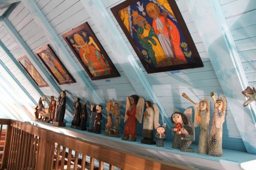 Fragment wystawy "Kultura Mazowsza w ludowej wizji świata". Ukośnie położony sufit, tuż pod nim półka, wszystko pomalowane na jasnobłękitny kolor. Na ścianie ludowe obrazy, na półce rzeźby - wszystkie dotyczą tematyki ludowej i przedstawiają świętych.
