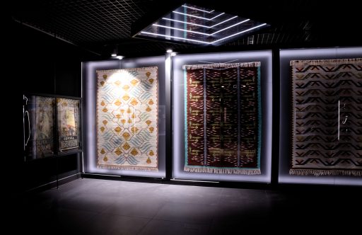Wystawa art deco w muzeum, ciemna sala, w niej na ścianie na wprost wiszą trzy duże szklane gabloty. W gablotach umieszczone są różnokolorowe kilimy w ludowe wzory.