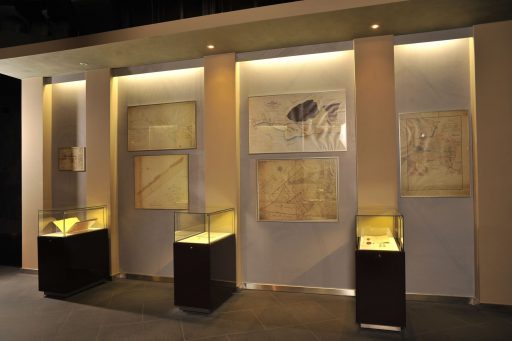Sala wystawy "X wieków Płocka" muzeum, na ścianach w szklanych ramach stare mapy, pod nimi w gablotach stare dokumenty.