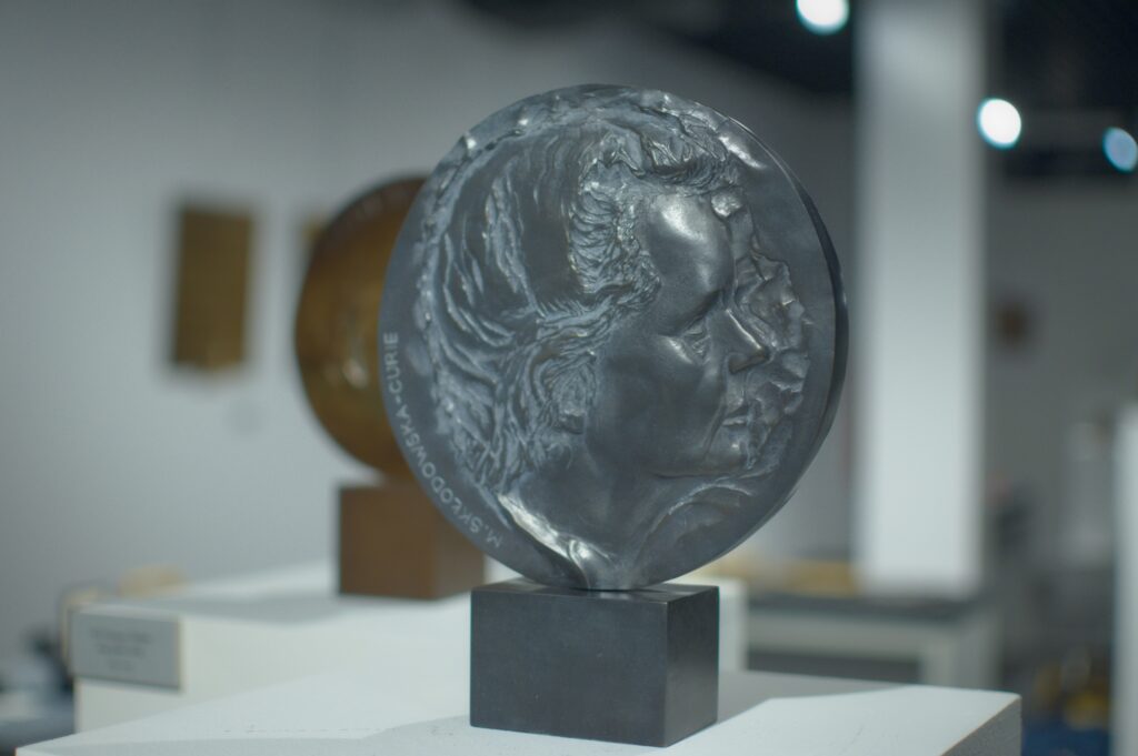 Medal z ciemnego metalu na srebrnej podstawce. Na medalu płaskorzeźba przedstawiająca twarz kobiety z profilu. To Maria Curie-Skłodowska.