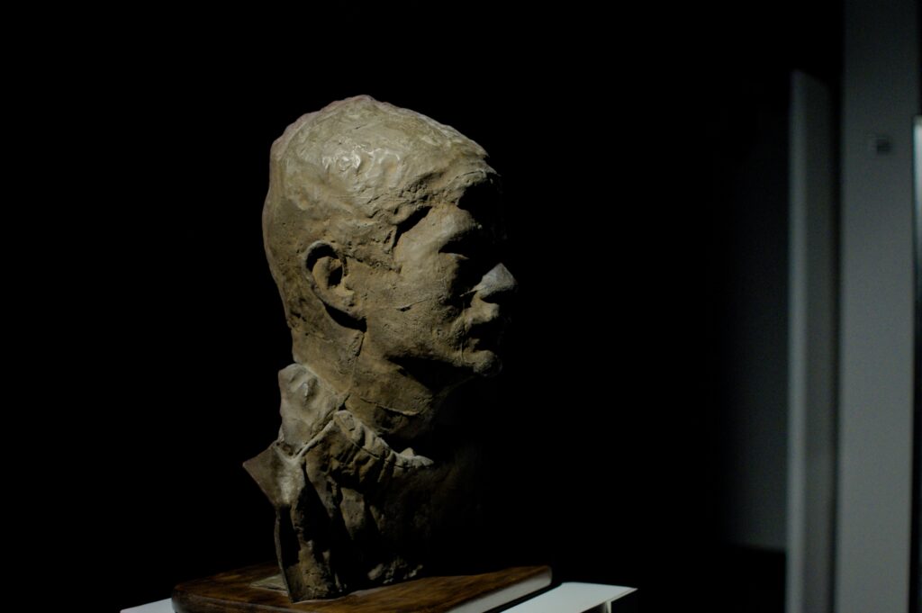 Rzeźba z gliny przedstawiająca głowę mężczyzny z wąsami.Rzeźba sfotografowana jest z profilu, na ciemnym tle. 