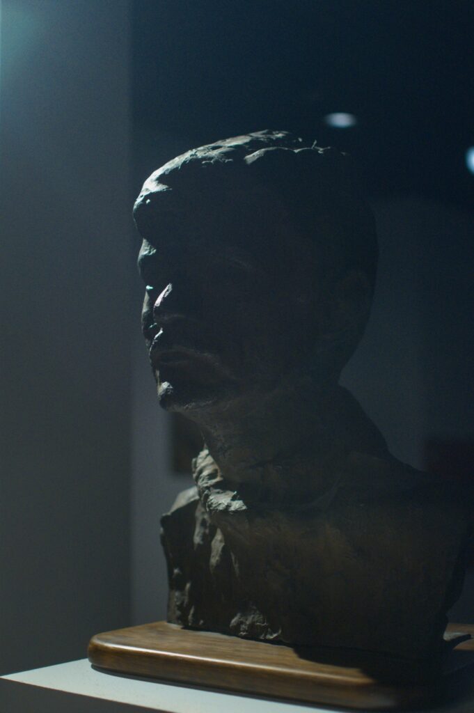 Rzeźba z gliny przedstawiająca głowę mężczyzny z wąsami.Rzeźba sfotografowana jest od przodu, na ciemnym tle. 