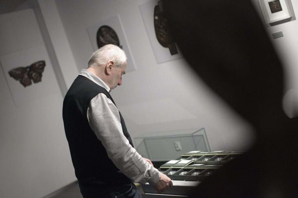 Wystawa rzeźb i medali w muzeum, starszy mężczyzna w ciemnej kamizelce ogląda medale umieszczone w niskiej, szklanej gablocie.