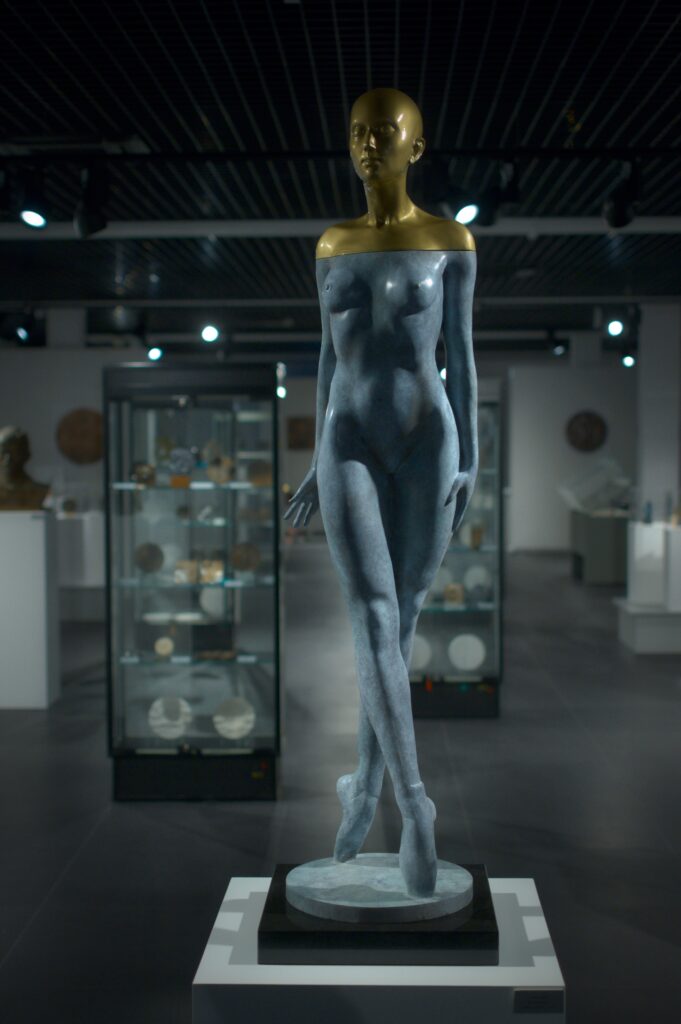 Wystawa rzeźby i medali w muzeum, w tle szklane gabloty z medalami, na pierwszym planie naturalnych rozmiarów rzeźba przedstawiająca nagą kobietę z łysą głową. Kobieta ma skrzyżowane nogi, na stopach ma baletki. Głowa i ramiona kobiety są z żółtego metalu, reszta jej postaci jest z szarego metalu.