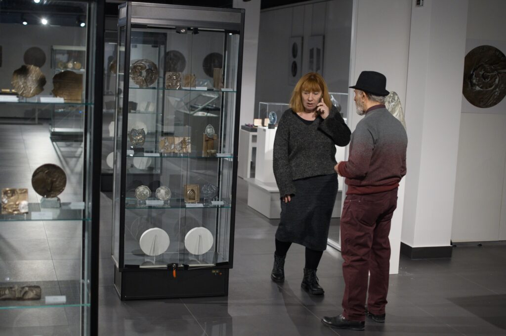 Wystawa rzeźby i medali w muzeum, po lewej stronie gablota z obiektami, po prawej stoją kobieta i mężczyzna, kobieta rozmawia przez telefon.