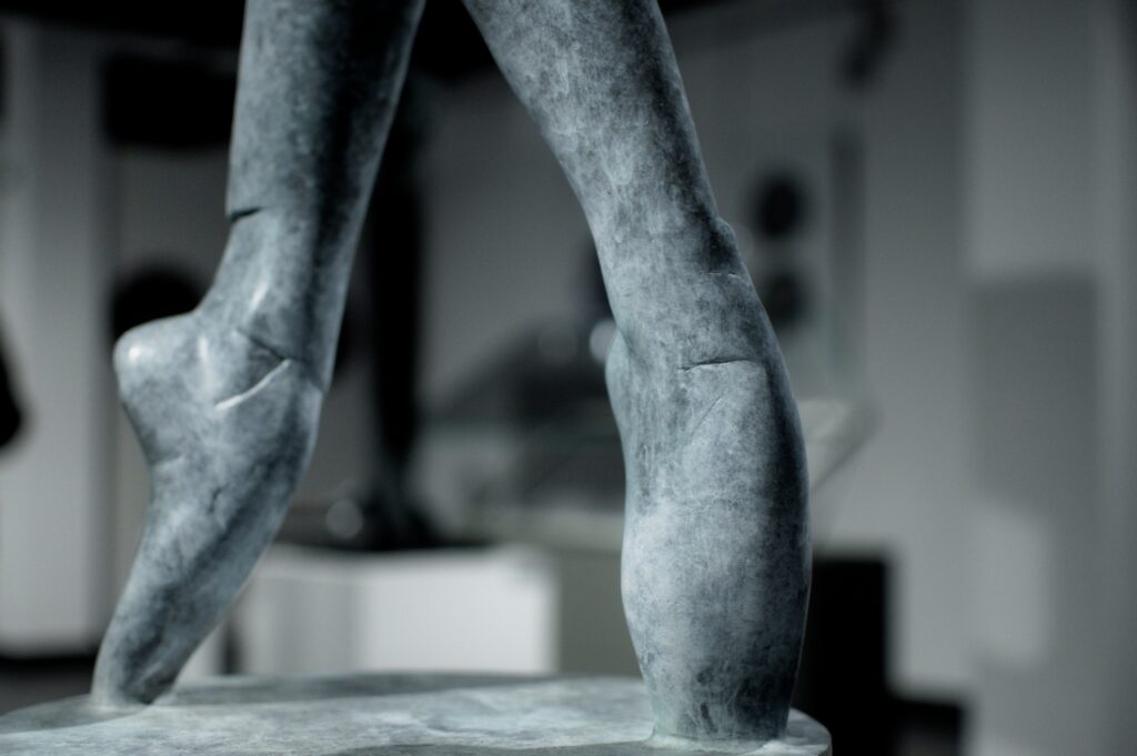 Zbliżenie na fragment rzeźby z szarego metalu, to stopy baletnicy w baletkacch stojące na czubkach butów.