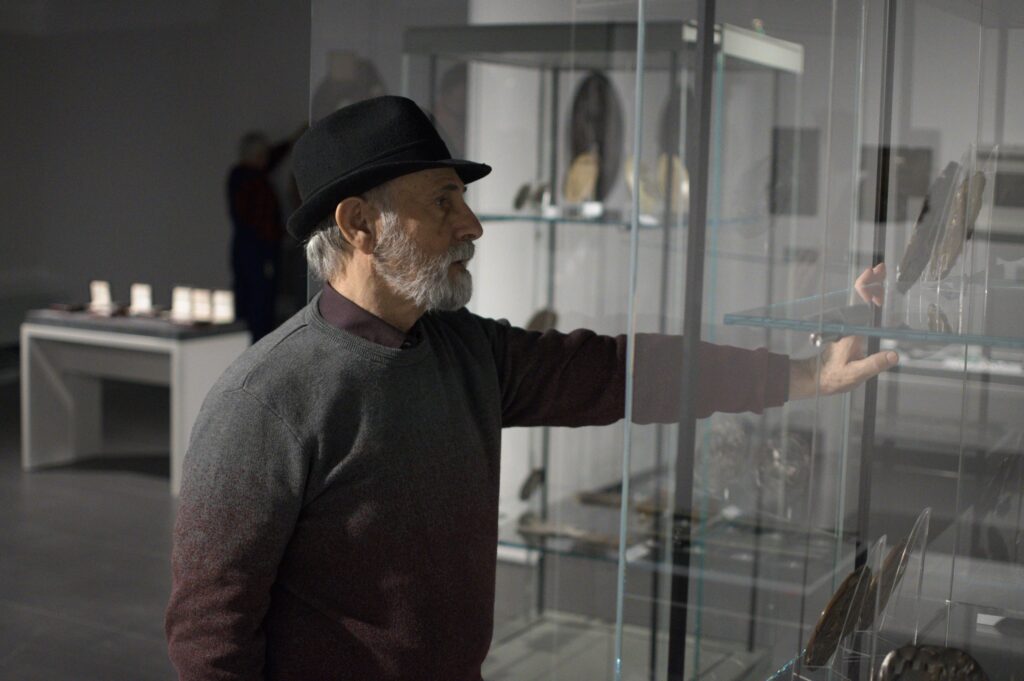 Wystawa rzeźb i medali w muzeum, przed szklaną gablotą z eksponatami stoi starszy mężczyzna z siwą brodą, w kapeluszu.