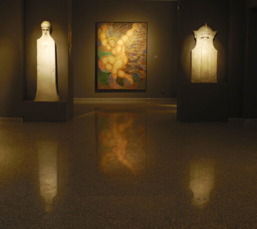 Wystawa Boesława Biegasa w muzeum, ciemna sala, na tle jednej ze ścian dwie rzeźby przedstawiające figury ludzkie, pośrodku duży, kolorowy obraz przedstawiający Tytana - nagiego mężczyznę w ekspresyjnej pozie. Na błyszczącej podłodze przed dziełami odbijają się ich wizerunki.