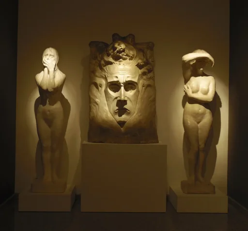 Wystawa Bolesława Biegasa w muzeum, ciemna sala, na tle ściany dwie białe, podświetlone rzeźby przedstawiające nagie figury ludzkie, jedna zasłania twarz dłońmi, druga podnosi ramię do twarzy, drugą ręką podtrzymuje piersi. Pośrodku duża płaskorzeźba przedstawiająca pociągłą twarz w ozdobnym ornamencie.