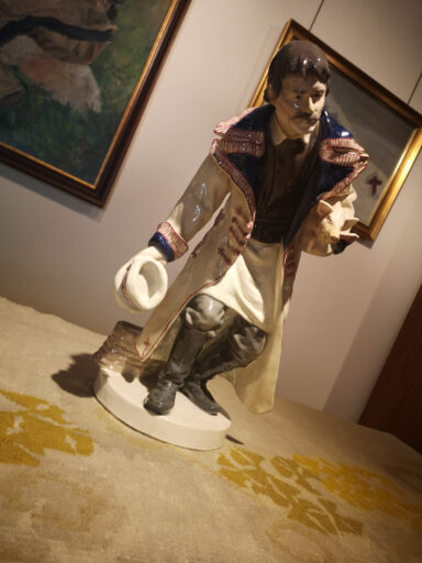 Sala ze sztuką secesyjną inspirowaną ludowością w muzeum. Na pierwszym planie stojąca na stoliku ceramiczna figurka mężczyzny w ludowym stroju, mężczyzna kłania się w pozie powitania.