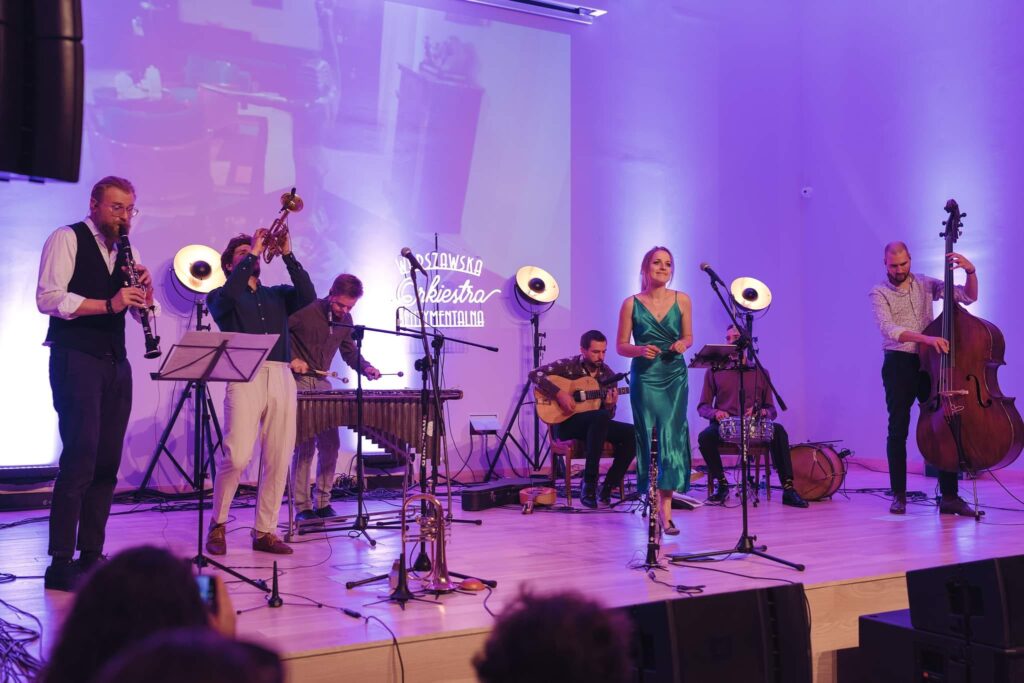 Zespół muzyczny na scenie muzealnej auli. Mężczyźni grają na różnych instrumentach, kobieta w zielonej sukni śpiewa do mikrofonu.