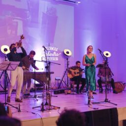 Zespół muzyczny na scenie muzealnej auli. Mężczyźni grają na różnych instrumentach, kobieta w zielonej sukni śpiewa do mikrofonu.