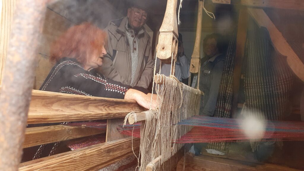 Dawny drewniany warsztat tkacki we wnętrzu drewnianej chaty, za nim rudowłosa kobieta objaśnia jego działanie stojącym obok trzem mężczyznom.