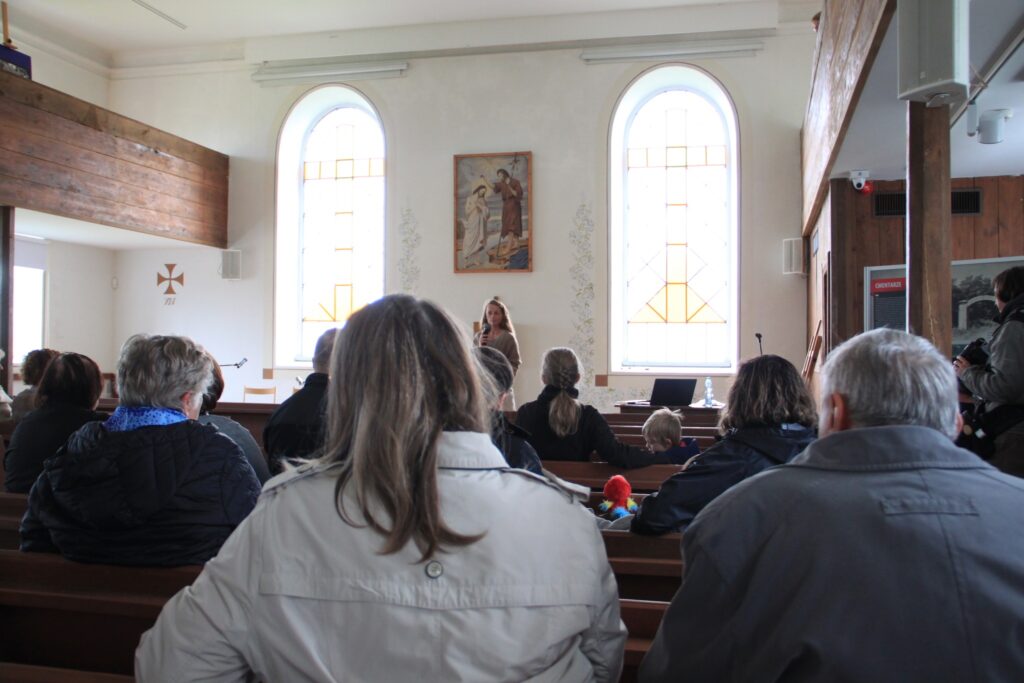 Wnętrze dawnego kościoła ewangelickiego w skansenie, w ławkach siedzą kobiety i mężczyźni w różnym wieku.