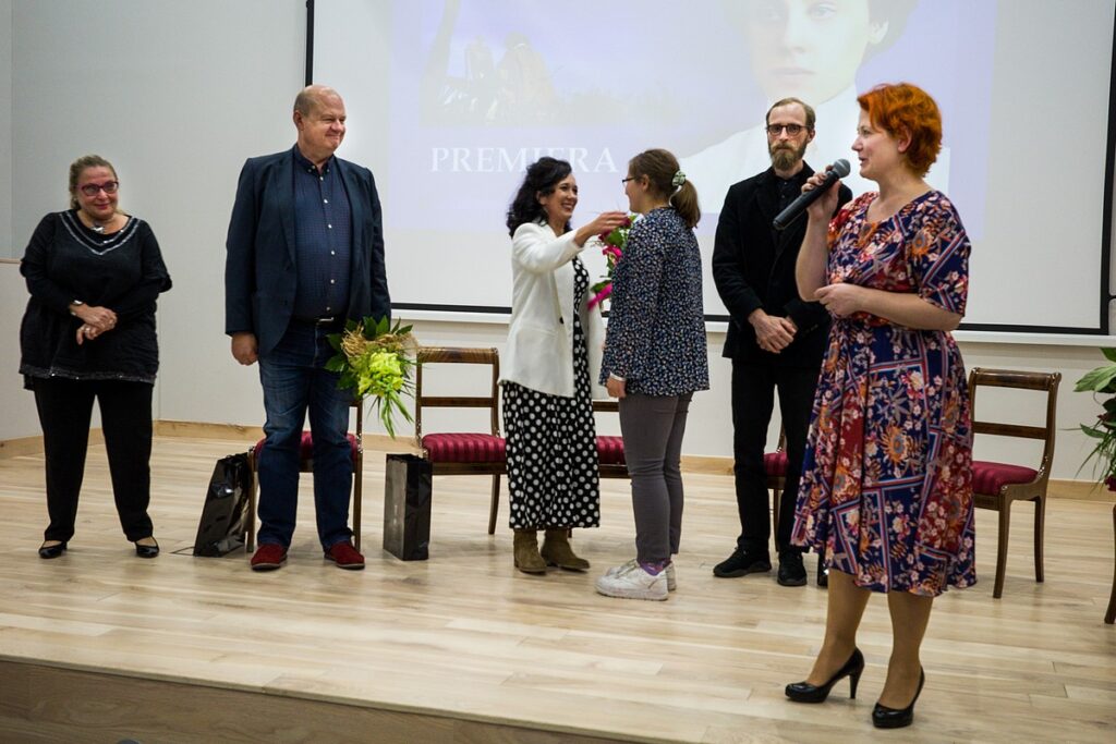 Grupa twórców filmu na scenie auli muzeum, na pierwszym planie, przy mikrofonie współautorka scenariusza Elżbieta Miecznikowska.  