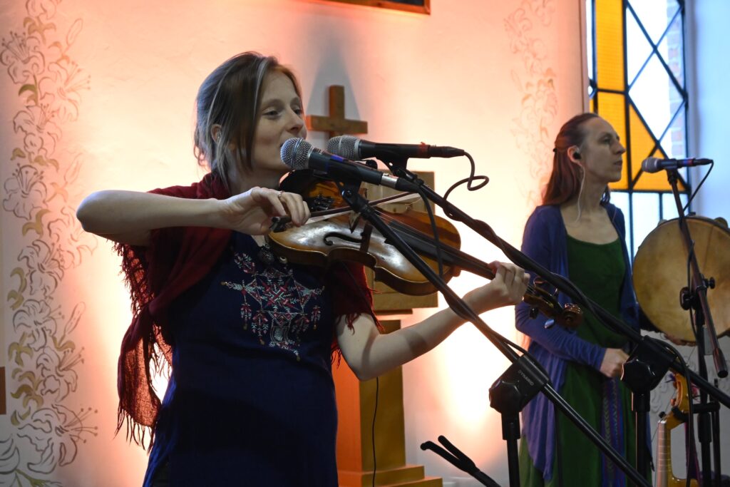 Zbliżenie młodej kobiety grającej na skrzypcach w pomieszczeniu. To członkini tria Sutari, które zagrało w skansenie. W tle widać powieszony na ścianie drewniany krzyż. 