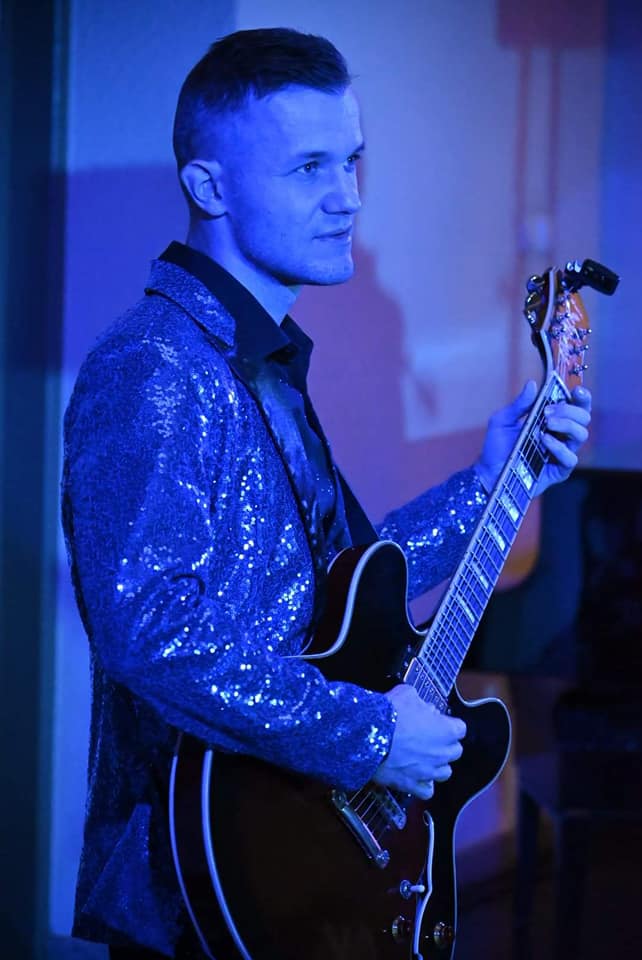 Młody mężczyzna w cekinowym garniturze gra na gitarze elektrycznej. Oświetla go niebieskie światło z reflektora. 