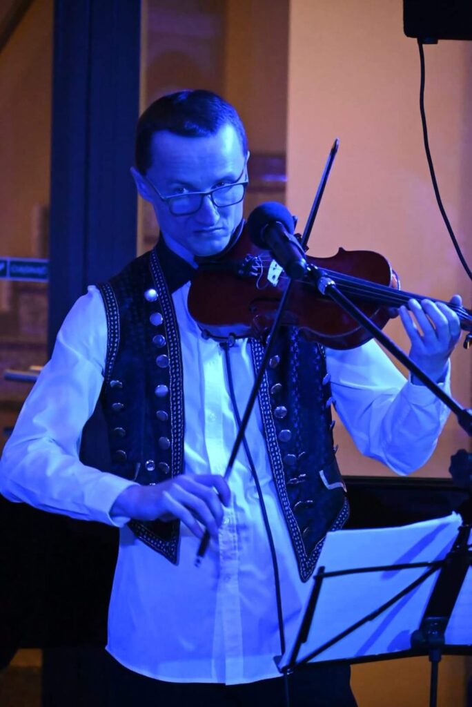 Młody mężczyzna w okularach, koszuli i kamizelce. Gra na skrzypcach, przed nim stoi mikrofon. Trwa koncert Bartosz Smorągiewicz Ensamble w Muzeum Mazowieckim w Płocku. 
