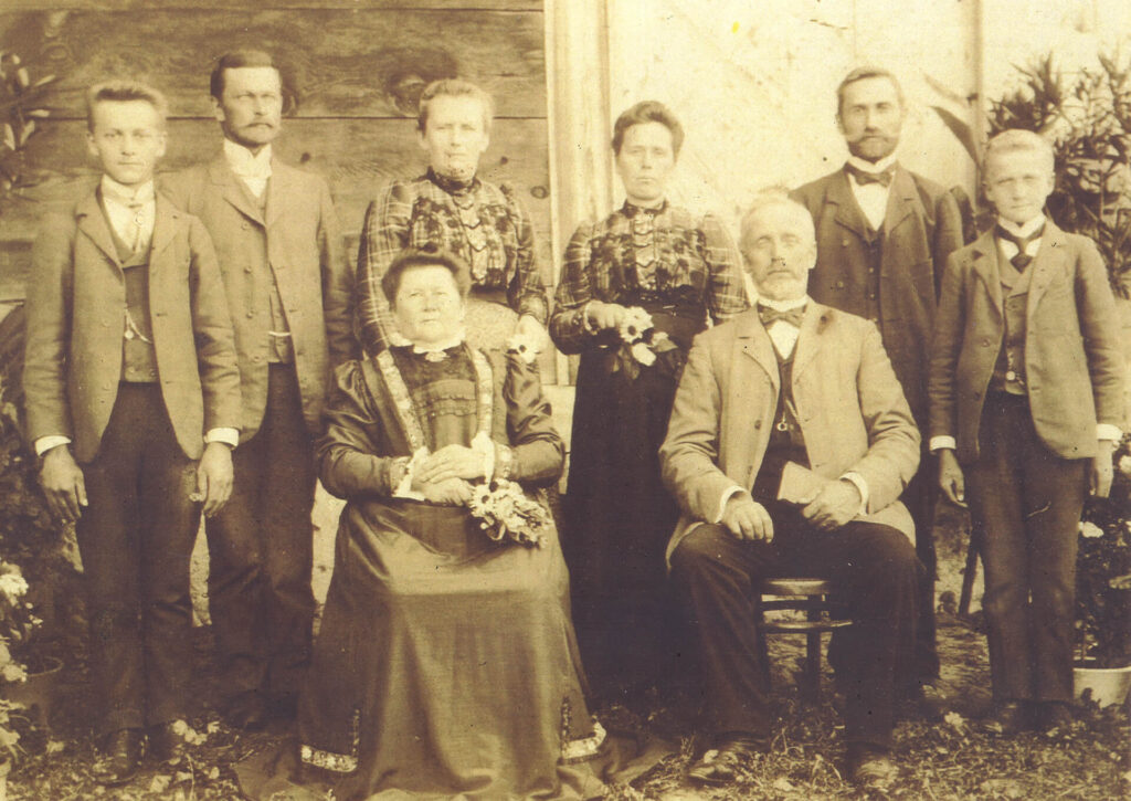 Archiwalna fotografia rodziny menonitów z XIX wieku. Czterech mężczyzn w surdutach, trzy kobiety w staromodnych sukniach i nastolatek są przed drewnianą chatą, najstarsza para siedzi.