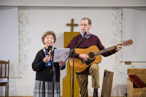 Za mikrofonem stoi starsza pani i mężczyzna z gitarą. Kobieta śpiewa piosenkę odczytywaną z kartki trzymanej w dłoni. Za nimi biała ściana, na ścianie wisi mały krzyż.