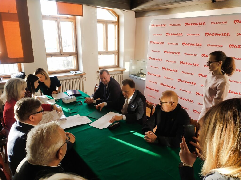 Grupa mężczyzn i kobiet siedzi przy stole przykrytym suknem. Za nimi bała ścianka z czerwonym napisem "Mazowsze". 