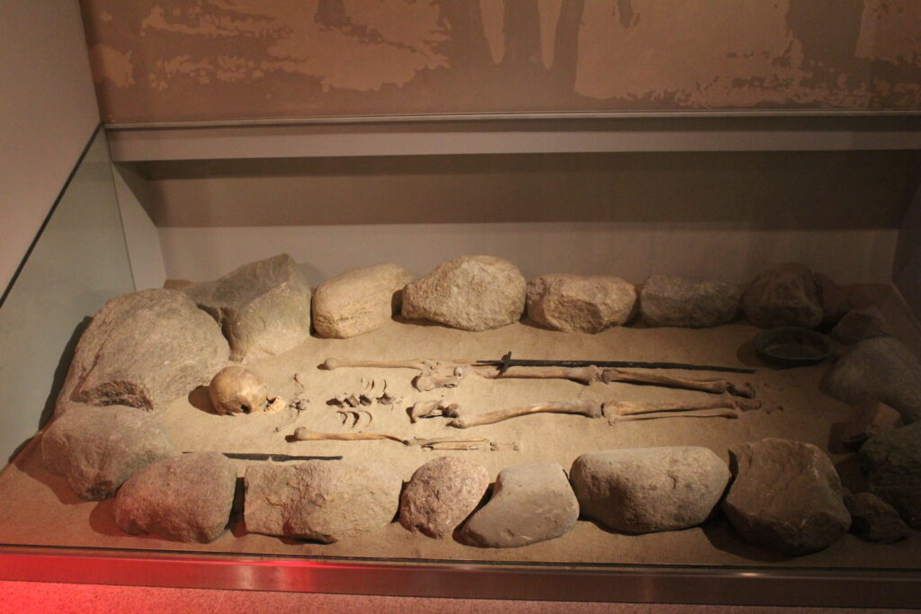Sala muzealna, w niej, na podłodze pod ścianą, pod szklaną gablota ludzki szkielet obłożony dookoła kamieniami. Obok szkieletu lezy miecz i misa.