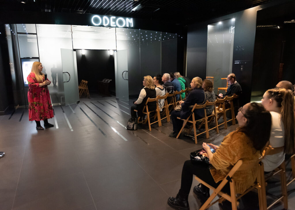 Muzealna sala, grupa osób siedzi na krzesełkach i słucha stojącej naprzeciw przewodniczki, młodej kobiety w sukni w kwiaty. Przed nimi wejście do małego kina, nad wejściem jasny napis "Odeon". 