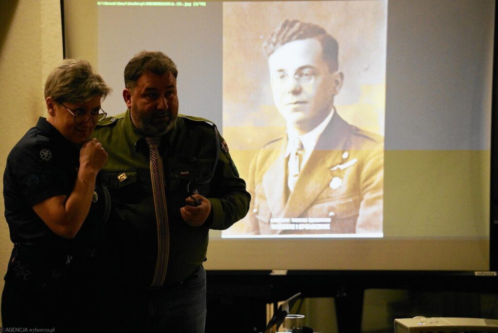 Kobieta i mężczyzna w harcerskich mundurach na tle ekranu z wyświetlonym na nim  czarno-białym zdjęciem Ładysława Żelazowskiego. To młody mężczyzna w okularach i w żołnierskim mundurze.