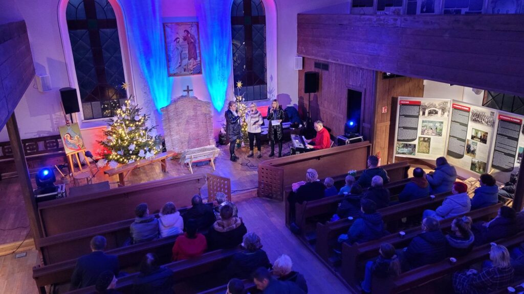 Jasełka w skansenie. Wnętrze małego, drewnianego kościoła sfotografowane z góry. Pośrodku, w miejscu ołtarza trzy kobiety śpiewają trzymając w dłoniach mikrofony, obok, za pianinem siedzi mężczyzna. Po drugiej stronie kobiet jest choinka. Naprzeciw są ławki, w ławkach siedzi publiczność. 