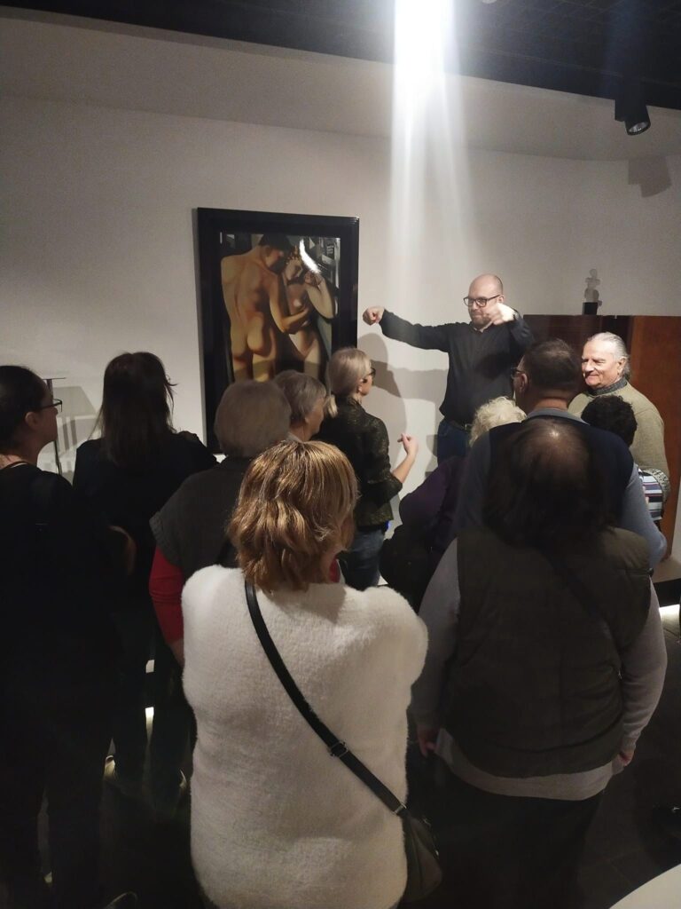 Jedna z sal wystawy art déco, Białe ściany, na ścianie na wprost patrzącego obraz przedstawiający nagie sylwetki mężczyzny i kobiety. Przed obrazem grupa zwiedzających, naprzeciw nich stoi gestykulujący mężczyzna w okularach. To tłumacz języka migowego.