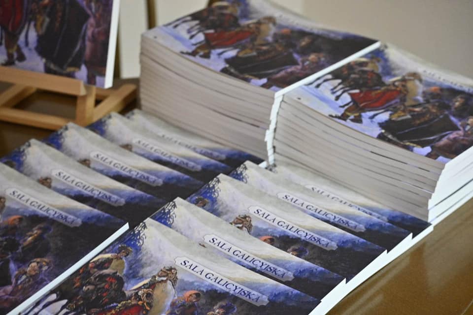 Ułożone w dwa stosy książki z napisem "Sala galicyjska" i kolorowym obrazem na okładce. Obraz przedstawia grupę osób w ludowych strojach na koniach na pokrytej śniegiem polanie.