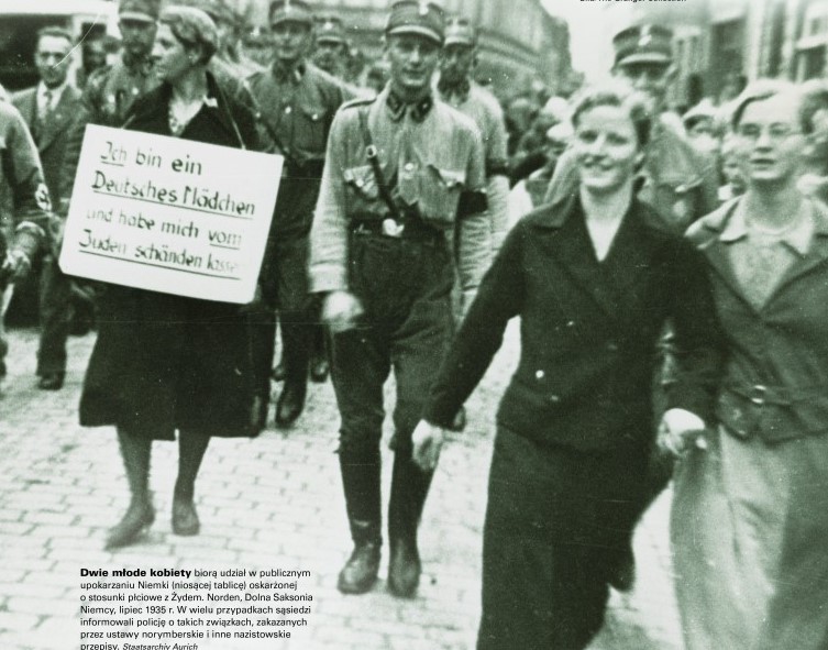 Archiwalne, czarno-białe zdjęcie z 1935 roku. Tłum ludzi w cywilnych ubraniach i mundurach idzie ulicą, po lewej stronie kobieta z kartonem na szyi. Na kartonie napis po niemiecku, informujący, że kobieta była w związku z Żydem. Po prawej stronie dwie inne młode kobiety śmieją się, patrząc w obiektyw. To publiczne upokarzanie Niemki podejrzanej o związek z Żydem.