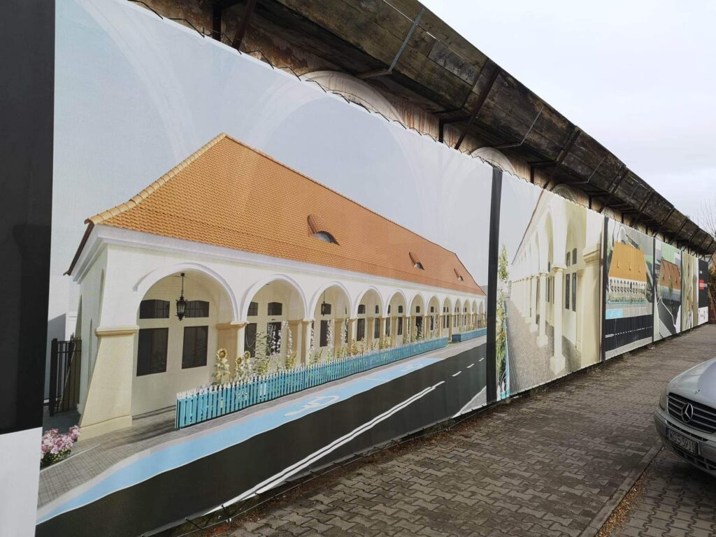 Budynek przyszłego muzeum w Gostyninie, jego fasadę zakrywa ogrodzenie z wydrukowaną na niej wizualizacją przyszłego wyglądu budynku. Przedstawia jasny parterowy gmach z czerwonym dachem i arkadami.