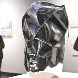 Wernisaż wystawy "Rekonfiguracje" w muzeum. Biała sala, na ścianach obrazy, w oddali dwie sylwetki zwiedzających kobiet. Na pierwszym planie szklana rzeźba ludzkiej głowy na postumencie, artystycznie przetworzona, przypominająca maskę w hełmie. Szło jest srebrne i przypomina metal.