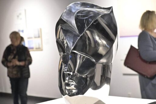 Wernisaż wystawy "Rekonfiguracje" w muzeum. Biała sala, na ścianach obrazy, w oddali dwie sylwetki zwiedzających kobiet. Na pierwszym planie szklana rzeźba ludzkiej głowy na postumencie, artystycznie przetworzona, przypominająca maskę w hełmie. Szło jest srebrne i przypomina metal.