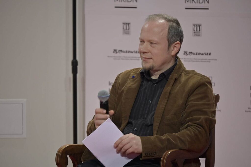 Konferencja otwierająca projekt "Opowieści". Scena muzeum, na krześle siedzi Andrzej Podściański, w dłoniach ma mikrofon i kartkę. 