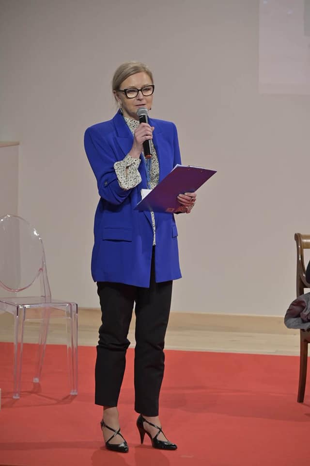 Aula muzeum, scena, na niej stoi Agnieszka Kaniewska-Płocieniak. Mówi do trzymanego w ręce mikrofonu.