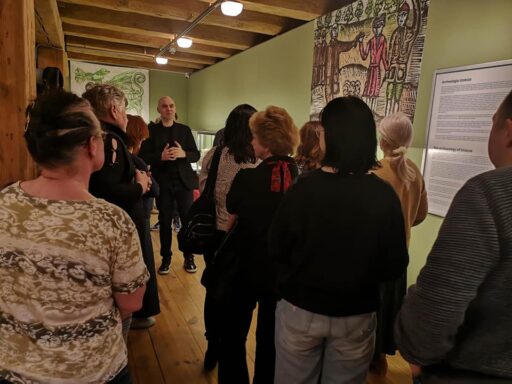 Wystawa "Nad Wisłą, na Urzeczu" w Spichlerzu. Po wystawie grupę zwiedzających oprowadza dr Łukasz Stanaszek. Wszyscy stoją przy zielonej ścianie, na której wisi duża reprodukcja ludowego rysunku.