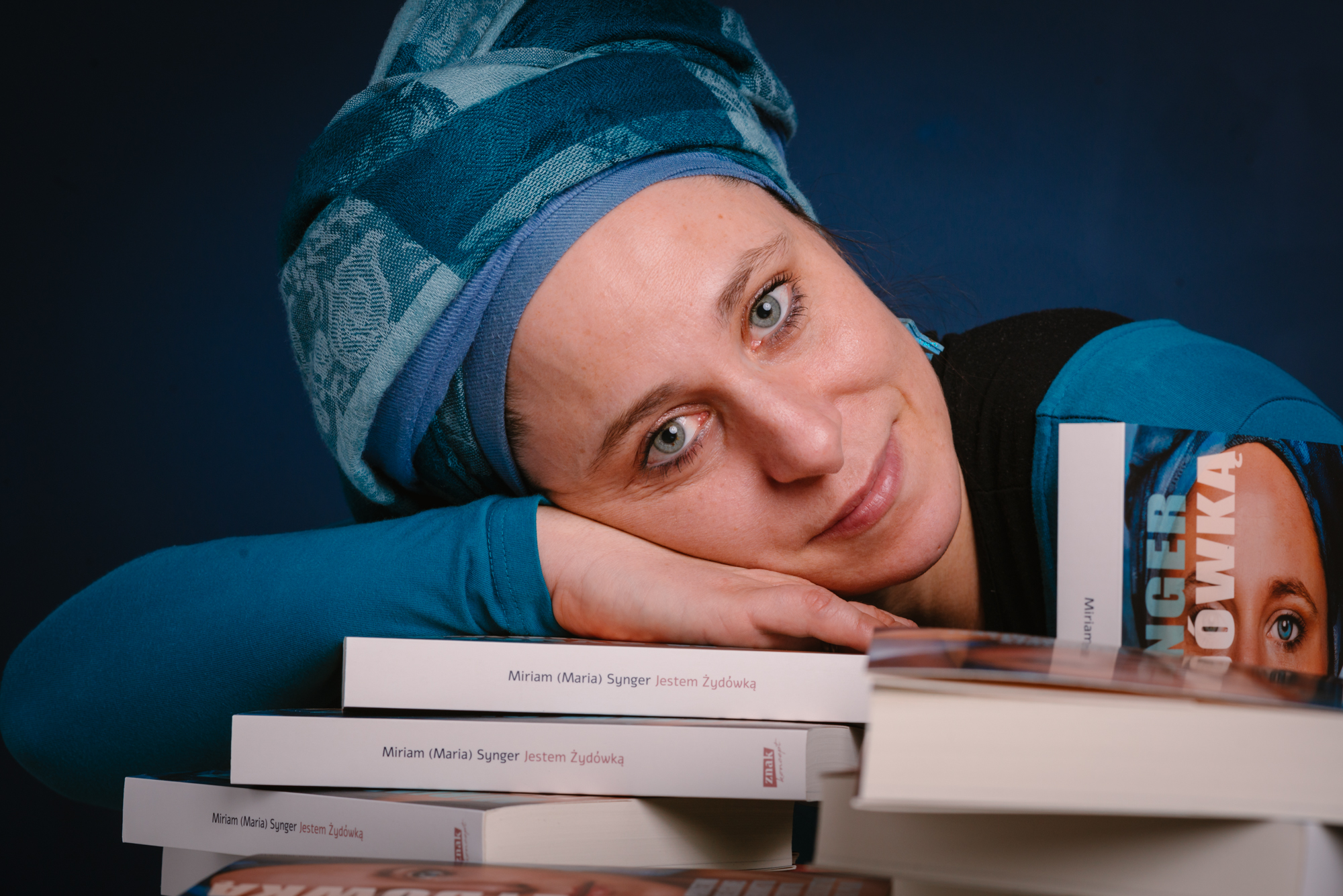 Miriam Synger, młoda kobieta z włosami ukrytymi pod zawojem, opiera dłoń i twarz na ułożonych przed nią książkach.